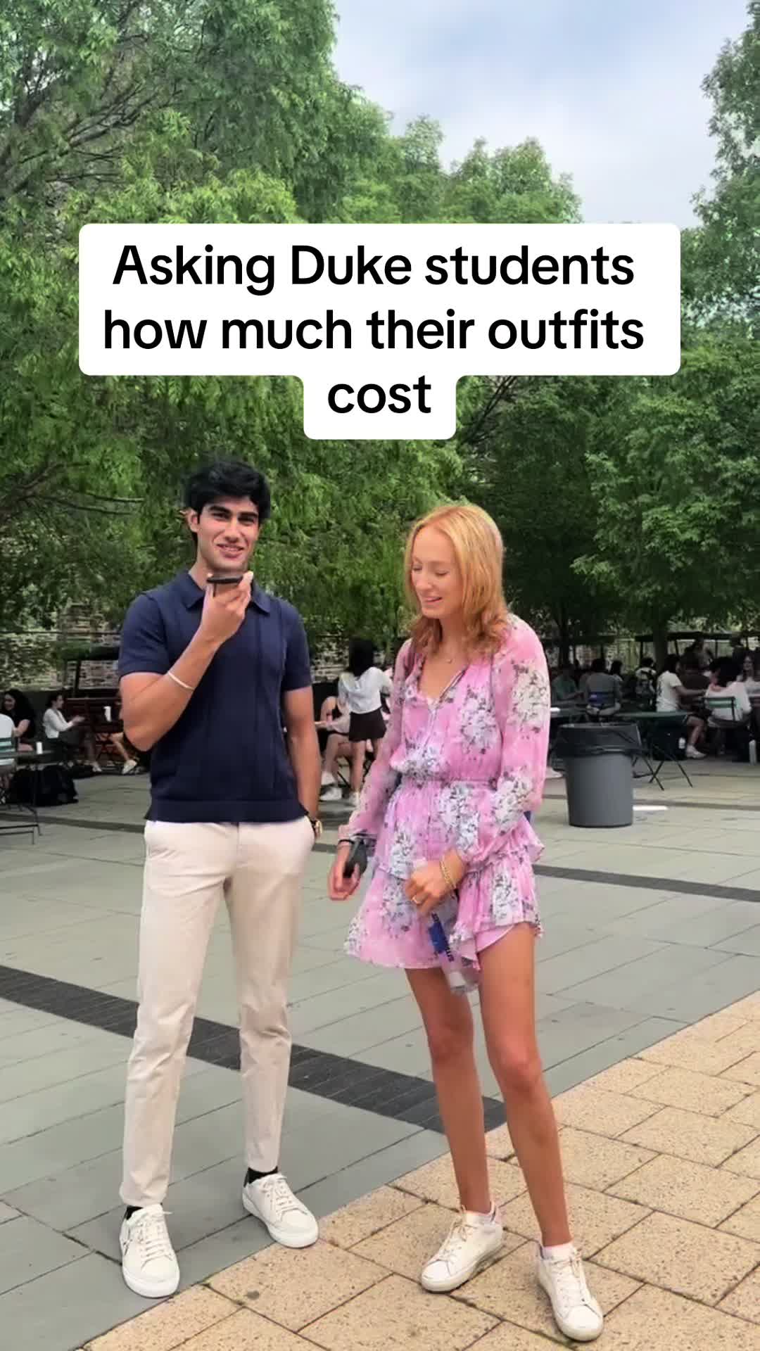 A street scene with two people, likely college students, being interviewed. A man with short dark hair and a navy polo shirt is holding a phone to his ear. A woman with long blonde hair and a pink floral dress is standing next to him, smiling. They are on a brick-paved area with other people and trees in the background.