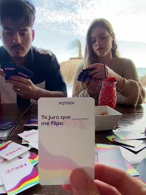 A group of people playing a card game at an outdoor table. A man and a woman face the camera, each handling cards. In the foreground, a card is held up displaying text. Background elements indicate a beach setting.