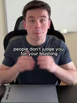 A medium shot of a man sitting in a chair, looking at the camera. The man is wearing a dark blue t-shirt. Above his head is a text overlay that reads, "people don't judge you for your blushing". The man is in a room, with a desk placed in front of him.