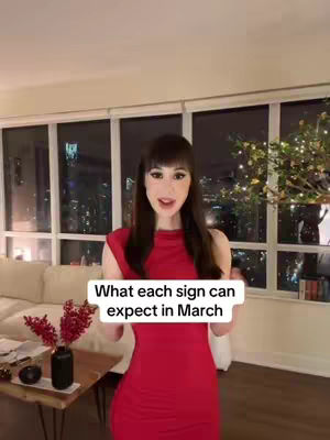 A woman in a red dress stands indoors, holding a sign with text. The background includes windows overlooking a city at night, a sofa, and decorative items.