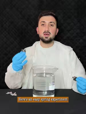 A man in a white lab coat and blue gloves stands in front of a beaker of water. A text overlay reads, 'Here's an easy spring experiment'. The background is a black surface.