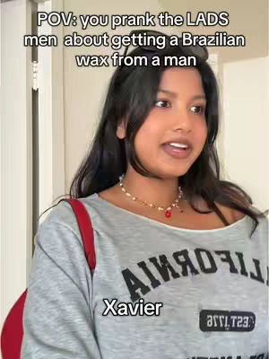 A woman with long black hair is in front of a white door. She's wearing a gray sweatshirt with the word "California" printed over it, alongside a red bag strap, and a gold necklace. Overlaid text says "POV: you prank the LADS men about getting a Brazilian wax from a man".