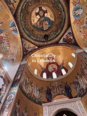 An interior shot of a church with ornate ceiling frescoes and artwork. The image is dominated by a circular painting of Jesus Christ at the top, with other biblical scenes and figures adorning the walls and ceiling. The composition is symmetrical, with intricate details and a golden color palette.