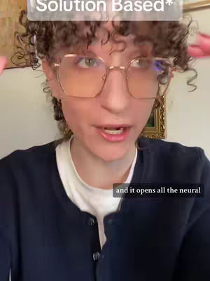 A woman with curly brown hair and square glasses looks directly at the camera while speaking. She wears a dark blue cardigan over a white top. The background includes a wall with a framed picture.