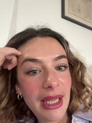 A close-up shot of a woman with light skin, light brown curly hair, and red lipstick. She's looking at the camera and touching her forehead with her right hand.