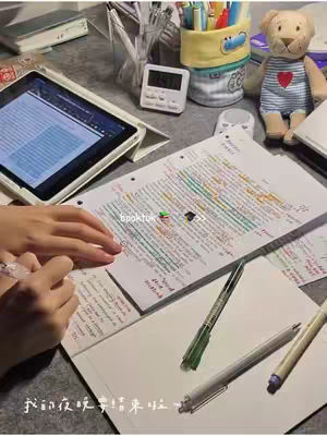 A study desk with various items: a tablet displaying notes, a notebook, pens, a timer, and a teddy bear. The scene is filled with items scattered across the desk. A pair of hands writing. The color palette is neutral with highlights from the writing and the items arranged.