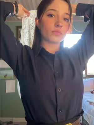 A young woman with long brown hair is posing with her arms raised above her head. She is wearing a dark blue button-down shirt. The background is softly lit.