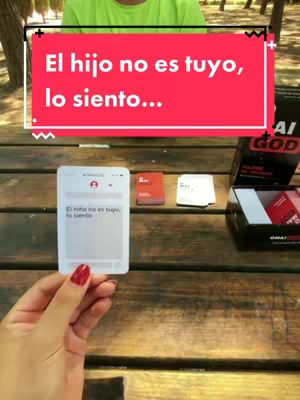 The image shows a table and a person. A person wearing a green shirt is in the background. The foreground has a wooden table. On the table, there's a game box with multiple cards and a big red text overlay. The text says, 'El hijo no es tuyo,
lo siento...' in the middle of the image .