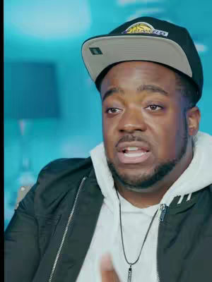 A medium shot features a Black man with a beard, wearing a Lakers baseball cap, black jacket over a white hoodie, and a necklace. He appears to be talking, with an open mouth and a slightly concerned expression. The background is a blurry teal color.