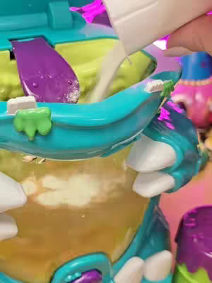 The image shows a close-up of a toy set up, viewed from above. A person's hand is pouring a white substance, possibly powder, into a container that looks like a toy. The container is multi-colored with shades of teal, yellow, purple, and white. The background has blurred, bright colors, primarily pink and purple, which suggests an interior setting.
