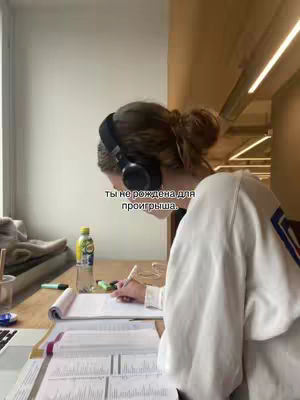 A woman, viewed from the side, is seated at a desk with books and papers. She is wearing a white sweater and headphones, with text overlayed on the image. The scene is lit by natural light coming from a window.