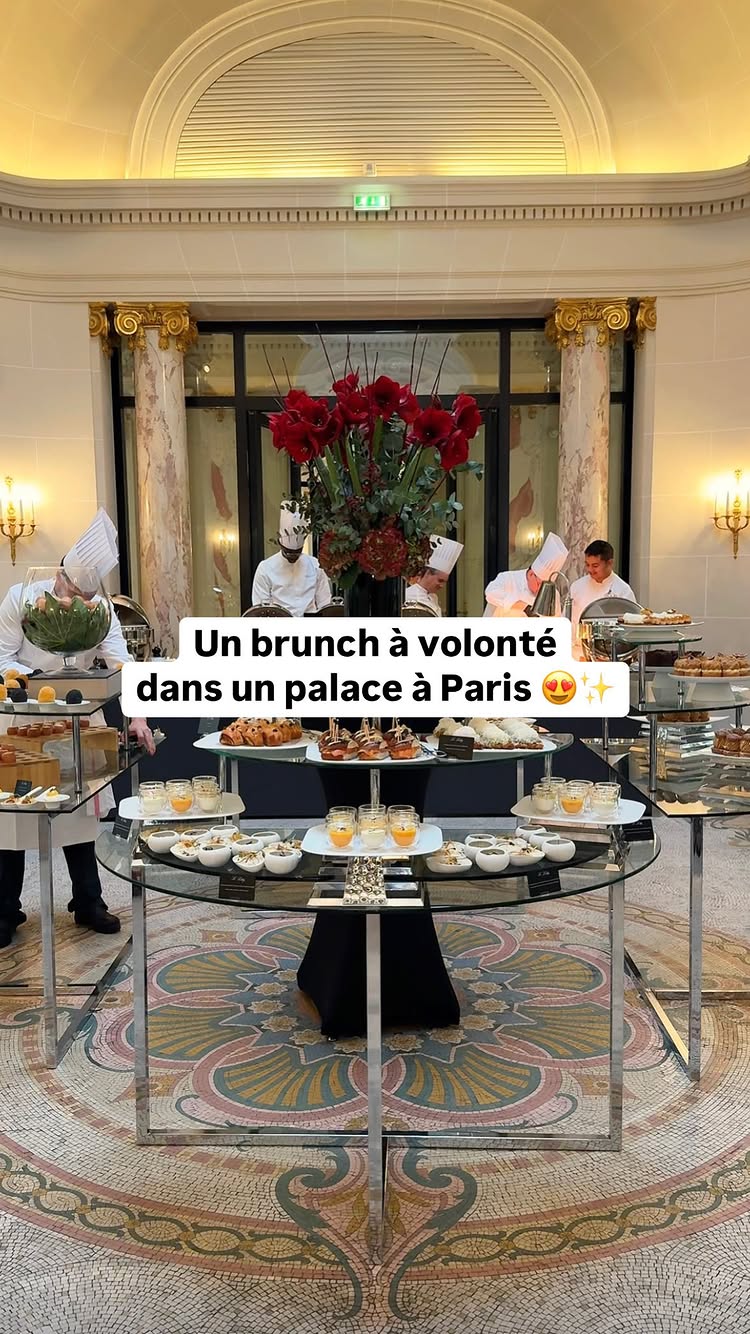 The image shows an elegant brunch setting inside a luxurious venue. In the foreground, there are glass-topped tables laden with a variety of dishes and drinks. Chefs in white uniforms are seen behind the food displays, preparing and presenting the food. The background features tall columns, architectural details, and floral decorations, suggesting a high-end restaurant or hotel.