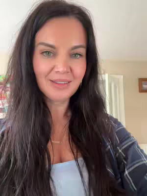 A woman with long dark hair looks directly at the camera. She is wearing a blue top and a dark plaid shirt. The background includes a room with beige walls and light-colored trim. The overall composition is a close-up, focusing on the woman's face and upper body.