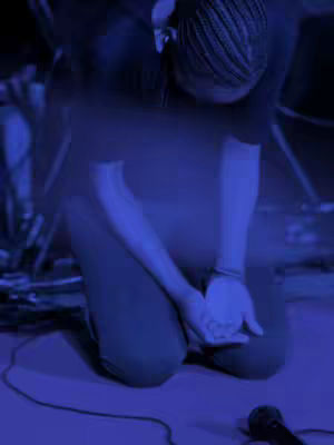 A person kneels on a stage with their head bowed downwards, hands clasped together, and a microphone in front of them. The color palette is dominated by a deep blue, creating a somber and focused atmosphere.
