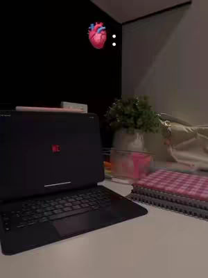 A close-up shot of a desk setup with a laptop displaying a red "MC" on the screen. A stylized heart emoji and two white dots float in the top left corner. Other elements include a plant, books, and a purse, all bathed in neutral lighting. The workspace is clean and organized.