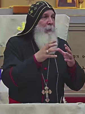 A man with a long white beard and a black hat speaks with his hands outstretched. He is wearing a dark suit. There is a crucifix hanging on a chain around his neck.