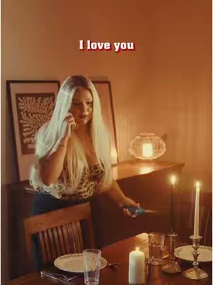 A woman with long blonde hair is on the phone in a warmly lit room, seemingly at a candle-lit dinner setup.
