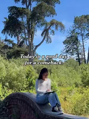 The image shows a young woman sitting on a stone bridge in a park-like setting. She is holding a pen and notebook, gazing down thoughtfully. The background has lush green trees and foliage, with a clear blue sky above. Text overlayed on the image states: 'Today is a good day for a comeback.'