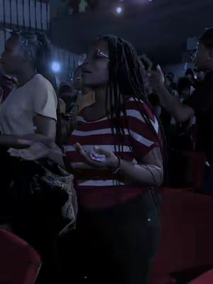 A dimly lit, interior shot focuses on a crowd, with a woman in the center who is wearing a red and white striped shirt. Several people are in the background, with some blurred and indistinct. The woman has black hair and appears to be in a moment of reverence, with her hands raised. The overall tone is reverent and spiritual, with a focus on the worship or gathering of a group.