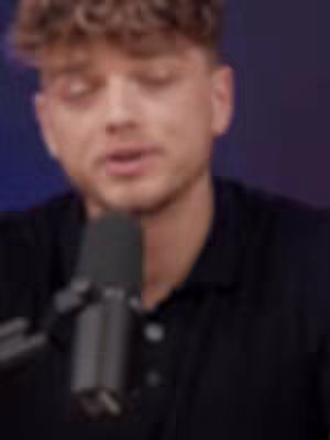 The cover image is a close-up shot of a man speaking into a microphone. He has short, textured hair and is wearing a black collared shirt. The background has a dark gradient of purples and blues. The microphone is in the center of the image. The overall composition is focused on the man's face and expression.