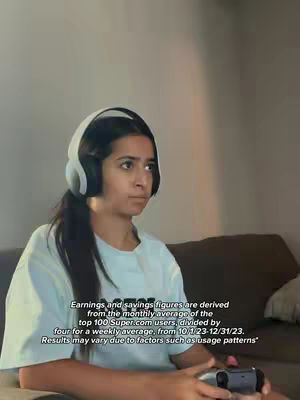 A young woman with long dark hair, wearing a white t-shirt and white headphones, sits on a brown couch while holding a game controller. The background is a neutral-toned wall.
