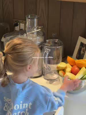 A young girl stands at a counter with her back to the camera, looking towards a bowl of fruit and a collection of kitchen appliances. The color palette is warm, with wooden cabinetry and beige countertops providing a neutral background. A text overlay reads 'building a home, not a rodeo'.