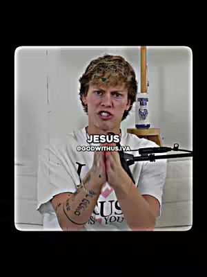 A young man with curly hair is centered, looking directly at the camera with a serious expression. He's wearing a white t-shirt and has a tattoo reading in hebrew on his left arm. Overlay text "JESUS" with a username is above his head. The background is a plain white wall and a blurred object.