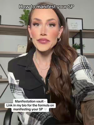 A woman with long brown hair and full makeup is centered in the frame. The top of the image has overlaid text, and below that is the woman's image. She is wearing a black jacket over a plaid shirt. Her expression is neutral with a slight smile. The background is a lightly blurred room with shelves and decor. The image has a soft, warm lighting.
