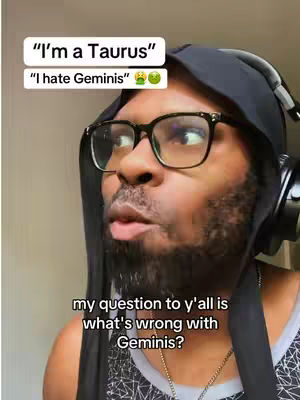 A selfie-style image of a Black man with glasses, a beard, and headphones around his neck. He is wearing a tank top and a black hood. The background is a plain, light color. Text overlays include 'I'm a Taurus', 'I hate Geminis', and additional text asking 'what's wrong with Geminis?'