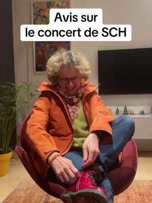 An older woman wearing glasses, an orange jacket, and blue jeans is seated in a red chair indoors. The background includes a framed picture, a flat screen TV, and some small decorative items. The text overlay reads "Avis sur le concert de SCH".