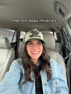 A woman wearing a baseball cap and denim jacket is sitting in the driver's seat of a car. She is smiling at the camera. The background shows the interior of the car, and the bright sun outside the front window.