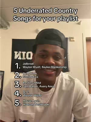 A man wearing a black cap is centered in the frame, smiling at the camera. Behind him, a black banner with stylized white letters is visible. To his left, a framed picture, partially cut off, adds to the background. The image features a list of country songs with their artists, overlayed in a white sans-serif font. The overall composition is a mix of personal and informative, suggesting a music recommendation video.