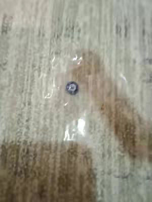 The image shows a partially transparent plastic wrapper on a surface. Inside, a small, circular metallic object with a dark center surrounded by a lighter ring is visible. The background appears to be a textured, light-colored surface, possibly fabric or paper, and the lighting seems soft and diffused.