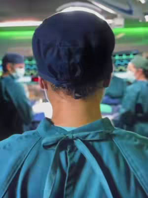 The image shows a person from the back, wearing surgical scrubs and a navy blue cap. The person is facing away from the camera with an operating room visible in the background. The scrubs are teal, and the background is blurred, with green lighting. There are other people, also wearing scrubs, in the background.