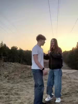 A couple stands facing each other outdoors, silhouetted against a setting sun and power lines. The woman is in a black bomber jacket and blue jeans, and the man in a white t-shirt and jeans. They appear to be embracing or about to embrace, with a background of trees and a muted sky.