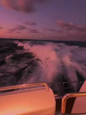 An eye level shot from a boat moving quickly through the water, creating foamy waves behind it. The water is dark, and the sky transitions from a vibrant pink near the horizon to a darker purple near the top. The boat's railing is visible in the foreground, with a sleek, rounded design.