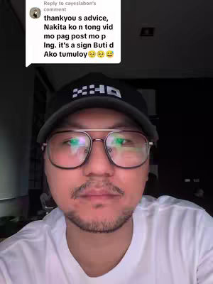 A selfie-style image of a man indoors, likely in a home setting. The focus is on the man's face, which is framed by large, round-framed glasses and a short, well-groomed mustache and goatee. He is wearing a dark cap and a white t-shirt. In the upper-left corner, a comment bubble contains overlaid text in a speech bubble, offering an intimate or casual impression.