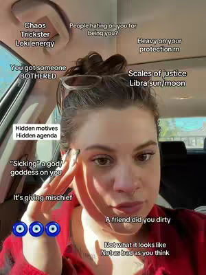 A selfie-style shot taken from inside a car featuring a woman in the foreground. She is looking directly at the camera, with her face and upper body visible. The background shows the car's interior, including the windshield and ceiling. She has brown hair, and is wearing glasses pushed up onto her head. Various text overlays are arranged around her head, providing astrological or psychic readings.