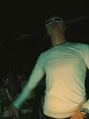 The image captures a man from a low angle, wearing white sunglasses, a white long-sleeve shirt, and a silver chain. Behind him, a dark background hints at a crowd.