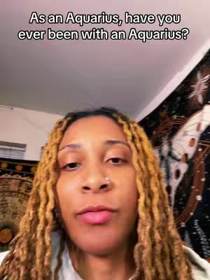 A selfie-style video with a woman in the foreground, focused from the mid-chest up, set against a backdrop that implies an indoor setting possibly in a room. The woman’s face is centered and a clear focal point. Her complexion is visible; she has brown skin. She has short, styled light brown or blonde hair, and a nose ring. The background displays an indistinct tapestry and a portion of a ceiling. The overall composition is well-lit, with the woman in clear focus.