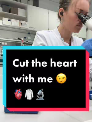 A person, likely a woman, in a white lab coat is looking into a microscope in a lab setting. The background includes lab equipment and supplies. The main focus is on text in a black rectangle that overlays the main image, containing the words: "Cut the heart with me 😉" with icons of a heart, a lab coat, and a microscope.
