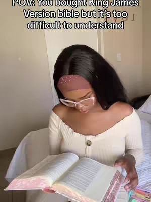 A woman with long black hair, wearing glasses and a white off-the-shoulder top, sits on a bed holding an open Bible. The image has overlaid text at the top that reads "POV: You bought King James Version bible but it's too difficult to understand."