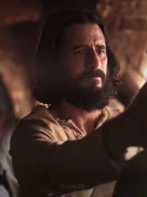 The image features a close-up shot of a man with dark, shoulder-length hair and a full, dark beard. He is looking slightly off-camera with a serious expression. He wears a light-colored garment, likely a robe or tunic, and the background is blurry with warm, muted tones.