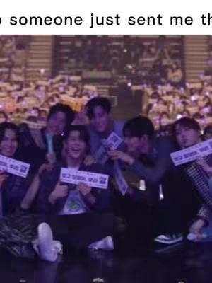 A group of young men, presumably a band or group of performers, are gathered together. They are holding up signs with Korean text, and appear to be on a stage or in a venue with a large audience in the background. The lighting suggests a concert setting with focus on the performers.