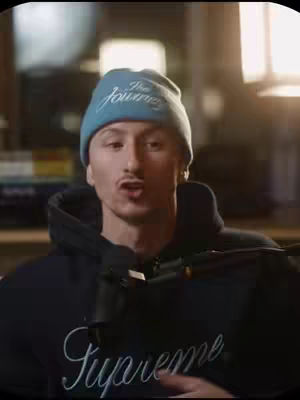A man with light skin, wearing a blue beanie with white text, a black hoodie with white script, and a microphone in front of him. The background is blurry but appears to be an inside setting. The man is speaking, looking directly at the camera.