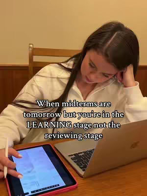 A young woman with long brown hair is sitting at a wooden table working on a laptop and an iPad. The laptop is open and the iPad is displaying what appears to be notes. The woman is leaning on her hand, looking down at the laptop and iPad, suggesting she is studying. Text overlayed on the image reads "When midterms are tomorrow but you're in the LEARNING stage not the reviewing stage".