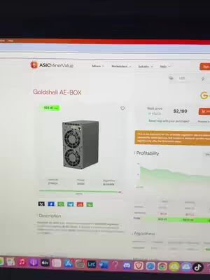 The image shows a computer screen displaying a website with a crypto mining device. The device is a grey box with two fans. The screen also displays price information and other details related to crypto mining.