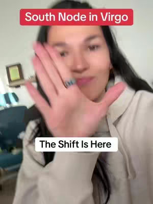A woman with long dark hair is holding up her hand towards the camera. The background is blurred, suggesting an indoor setting. Two text overlays are present: 'South Node in Virgo' in a red rectangular box at the top and 'The Shift Is Here' in a white rectangular box at the bottom. She is wearing a light cream colored jacket, and a ring in her finger.
