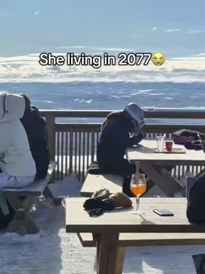 A person is sitting at a table on a deck overlooking a snowy landscape. The person is leaning over towards the table like something is stressing them out, and there is a drink and phone sitting in front of them.