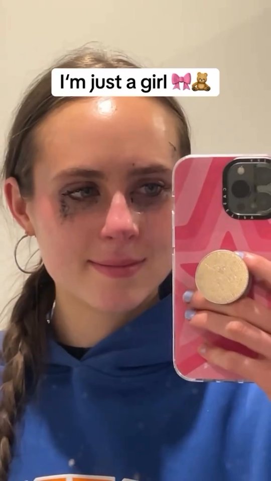 A selfie-style shot showing a young woman looking into a mirror, likely in a bathroom. Her face is framed by her hair, which is pulled back in a braid. The main subject has tears, and her mascara is smeared, suggesting she has been crying. The background includes a white wall and part of a mirror, in which the woman's phone is held. A text overlay says, 'I'm just a girl